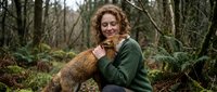 Young Caucasian woman with curly hair embracing red fox in mossy forest setting. Wildlife conservation and nature connection concept for outdoor campaigns.