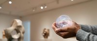 Person holding transparent crystal sphere in modern art gallery with sculptures and warm lighting creating contemplative museum atmosphere.