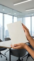 Person with medium skin tone holding blank white notebook in modern office with floor-to-ceiling windows and city skyline view, suitable for business, education, and corporate branding projects.