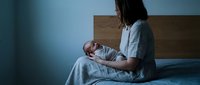 Young Asian mother in white pajamas holding sleeping newborn baby while sitting on bed in dimly lit bedroom during nighttime feeding routine.