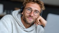 Young bearded Caucasian man in glasses and headphones smiling while working on laptop in modern office environment for remote work and online communication.