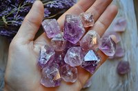 Raw amethyst crystal chunks displayed in open palm with lavender flowers in background, showcasing natural purple gemstone minerals for spiritual healing.