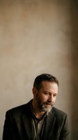 Middle-aged man with dark hair and full beard wearing olive blazer and neutral shirt, looking downward against warm beige wall backdrop for portrait and professional use.