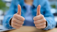 Close up view of hands showing thumbs up gesture against blurred blue clothing background, expressing positivity and approval in workplace setting.