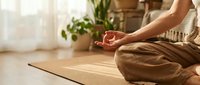 Woman in casual beige clothing meditating on yoga mat indoors with potted plants and natural light, wellness and mindfulness practice.