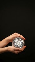 Two hands holding a glowing crystal ball with swirling light energy inside against dark background. Suitable for mysticism, fortune telling, and spiritual concepts.