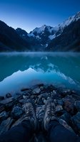 Hiker's boots resting on rocky shore of turquoise alpine lake with snow-capped mountain peaks reflected in calm water at dawn, adventure and travel concept.