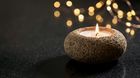 Glowing candle in textured stone holder creates warm ambiance with golden bokeh lights background for relaxation and meditation atmosphere.