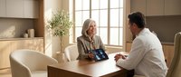 Senior Caucasian woman consulting with male doctor about medical results on tablet in modern clinic office with natural lighting and minimalist design.
