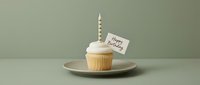 Minimalist birthday cupcake with white frosting, striped candle, and happy birthday card on a sage green background for celebration concepts.