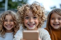 Cheerful curly-haired children laughing while looking at smartphone screen together. Close up portrait of happy kids sharing digital device outdoors.