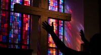 Silhouette of person with raised hands in worship before wooden cross with colorful stained glass window light creating spiritual atmosphere for faith content.