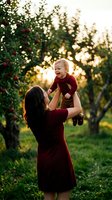 Mother lifting happy toddler in apple orchard during golden hour sunset creating warm family bonding moment in nature with soft bokeh background.