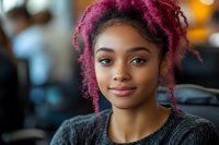 Young African American woman with vibrant magenta curly hair smiling confidently at camera, wearing black sweater against soft bokeh background. Natural makeup and warm expression.