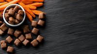 Dry pet food kibbles scattered on dark wooden background with fresh carrots. Bowl of natural dog treats for healthy nutrition. Copy space for text.