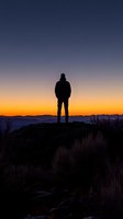 Silhouette of person standing on mountain peak at sunset, dramatic orange and purple sky backdrop with mountain ranges and dry grass in foreground.