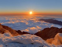 Dramatic sunrise over sea of clouds from snowy mountain peak, golden sunburst illuminating fluffy cloudscape, rocky mountain ridges emerging through misty morning atmosphere.