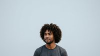 Young man with curly dark hair and medium brown skin tone wearing casual gray t-shirt, smiling against neutral white background for portrait and headshot use.