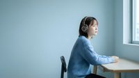 Young East Asian woman wearing headphones, seated at desk by window in minimalist office space, listening to music or audio content during work break.