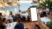 Hand of a man with dark skin tone holding a smartphone with blank white screen at outdoor dining event with blurred guests and coastal scenery in background.