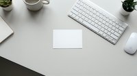 Clean white desk workspace with blank business card mockup, keyboard, mouse, coffee cup and plants for branding design projects.