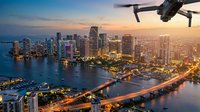 Drone flying over illuminated downtown skyline at sunset with waterfront, high-rise buildings, marina, and glowing city lights reflecting on water below.