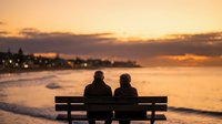 Elderly couple sitting on bench watching romantic sunset over ocean beach with golden sky and gentle waves creating peaceful retirement moment.