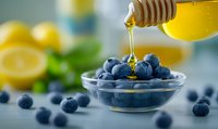 Fresh blueberries in glass bowl with honey drizzling from wooden dipper, fresh citrus fruits and mint leaves in soft focus background, healthy breakfast concept.