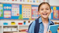 Happy elementary school girl with backpack holding books in colorful classroom ready for learning and education activities.