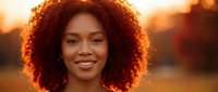 Young African American woman with curly hair smiling warmly during golden hour sunset, natural beauty portrait with soft bokeh background lighting.