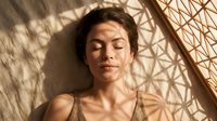 Young Asian woman relaxing with eyes closed in warm sunlight with geometric shadow patterns on face and body creating peaceful spa wellness moment.