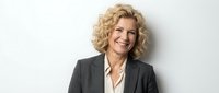 Professional blonde businesswoman in dark suit smiling confidently against white background, corporate headshot for business presentations and marketing.