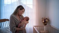 Young Caucasian mother in cozy sweater tenderly holding newborn baby in bright nursery room with wooden crib and soft natural lighting from window.