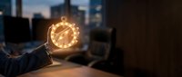 Businessman hand holding glowing clock icon in modern office at dusk representing time management and productivity concepts for business success.