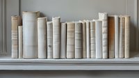 Row of vintage books in white and beige tones arranged on wooden shelf against neutral wall, creating minimalist monochromatic library composition.