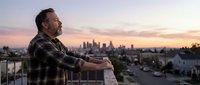 Middle-aged Caucasian man in plaid shirt contemplating city skyline at sunset from balcony, urban lifestyle and reflection concept for wellness content.