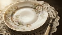 Vintage porcelain plate with delicate floral rose design and ornate rim, styled on lace doily with antique fork for elegant table setting and dining decor.