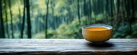 Ceramic bowl with golden tea on rustic wooden table overlooking serene bamboo forest, creating tranquil zen atmosphere for meditation and relaxation.