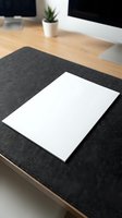 Blank white paper sheet on dark desk mat in modern office workspace with computer monitor and plant for business presentations and documents.
