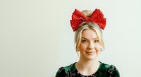 Young woman with blonde hair and fair complexion wearing a red sequined bow headband and dark green sequined top, smiling at camera against neutral background. Suitable for holiday, festive, fashion, and portrait design projects.