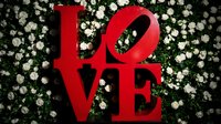 Red LOVE sign sculpture against a lush white rose flower wall backdrop, romantic decor for weddings, Valentine's Day, and events.