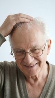 Senior man with white hair and glasses touching his head, smiling while experiencing hair loss, suitable for healthcare, wellness, and medical content.