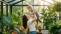 Young Caucasian mother lifting happy toddler in a lush greenhouse, surrounded by tropical plants, celebrating family love and nature.