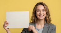 Young Caucasian businesswoman in gray blazer holding blank white paper sign with copy space, smiling at camera against yellow studio background.