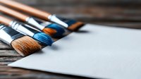Professional art brushes with blue and brown bristles lying on rustic wooden surface next to blank white paper, ready for creative painting session.