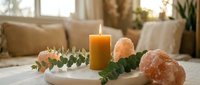 Cozy home spa setup with burning candle, eucalyptus branches and himalayan salt stones on white table in bright living room for wellness relaxation.
