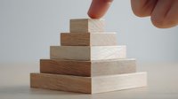 Finger placing final wooden block on top of pyramid structure, demonstrating hierarchy, growth, and achievement in business concept.