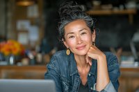 Confident mature Asian woman with gray hair smiling in cafe while working on laptop, wearing denim shirt and gold earrings.