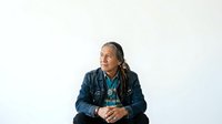 Middle-aged man with long dark hair and Native American ethnicity wearing layered denim and blue shirt, sitting with hands clasped, isolated on white background for portrait and character design use.
