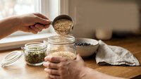Hand pouring quinoa seeds from wooden spoon into glass jar for healthy meal prep and pantry organization in modern kitchen setting.